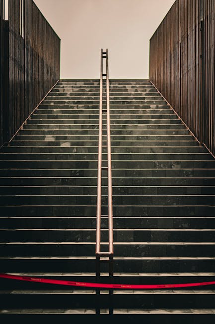 Black and white photograph of an external fire escape staircase made of metal, featuring multiple landings with white railings and grated metal steps, attached to the side of a building with concrete